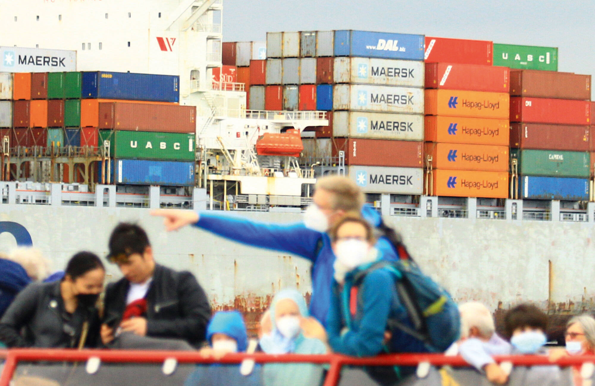 Passanten auf einer Fähre der HADAG Seetouristik und Fährdienst AG. Im Hintergrund wird ein Containerschiff entladen. Foto: IMAGO / Hanno Bode